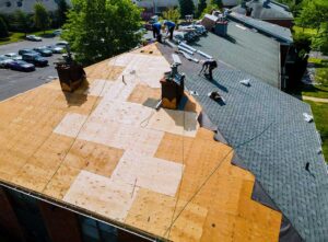 A roof installer works on a home, adding modern shingles with snow on the ground and trees in the background.
